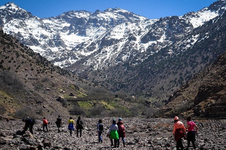 Hikers trekking through scenic trails in the High Atlas Mountains with snow-capped peaks, green valleys, and traditional Berber villages in the background