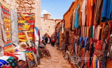 Colorful local market in Ait Ben Haddou showcasing handmade Moroccan rugs, textiles, and crafts during a day trip from Marrakech to Ouarzazate.